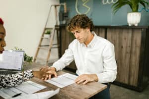 A man and woman are sitting at a business bank desk discussing the difference between profit and profitability. The business bank executive explains to the man why profitability is the key to making his business loan-ready.