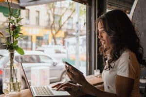 A woman is sitting at a table, using a laptop and cell phone, looking for information about how non-dilutive funding can benefit her businesses.