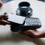 An image of a person with a phone tapping to pay at a restaurant represents the financial aspects of a business line of credit and it’s costs.